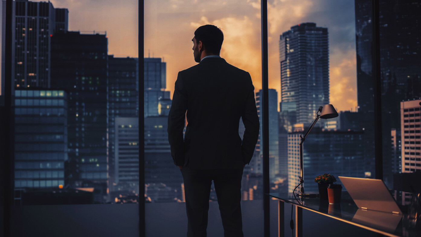 Businessman standing alone in a high-rise office overlooking a city skyline at night, symbolizing ambition, achievement, and modern performance culture.
