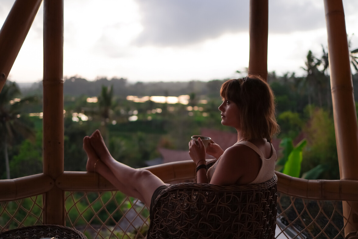 Woman resting quietly on a balcony with a warm drink, reflecting while overlooking a peaceful natural landscape.