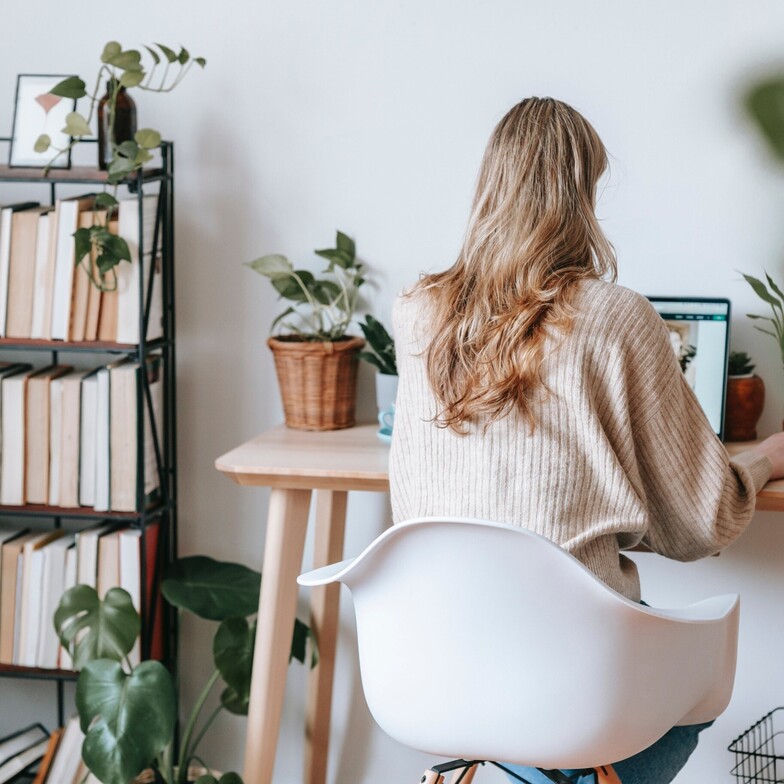 A woman seated at a desk using a laptop, surrounded by books and houseplants.